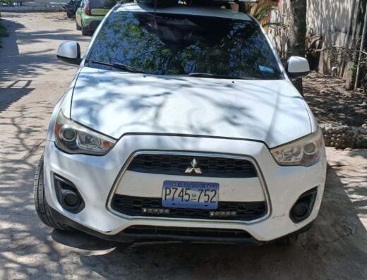 White Mitsubishi Outlander parked outdoors with roof cargo box, surrounded by trees and a rustic setting, under a clear sky.
