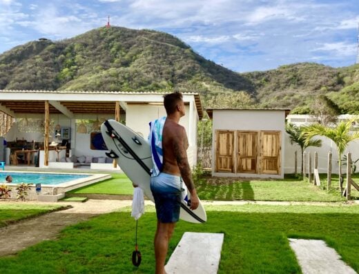 Man holding a surfboard in a lush garden with mountains in the background at Villas San Lorenzo Surf & Stay. Perfect for surfing classes and tours in a scenic coastal setting.