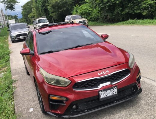 Red Kia Forte parked on the side of a road with greenery and other vehicles in the background, under a cloudy sky.
