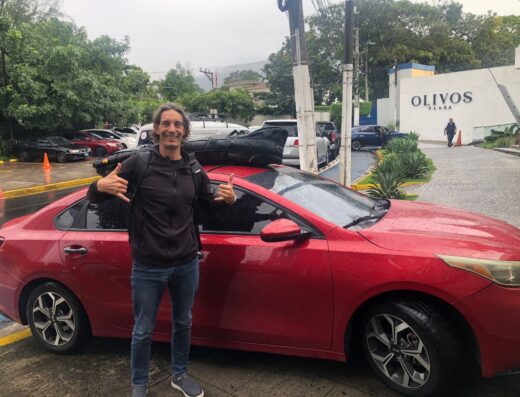 Ricardo López standing next to a red car with surf gear on top, in El Salvador, promoting surf taxi services for surfers.