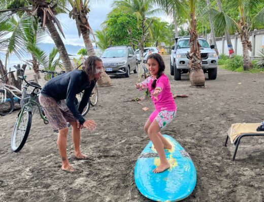 Child surfing on a blue surfboard with an adult nearby, surrounded by lush palm trees and parked cars in Jacó, Costa Rica.