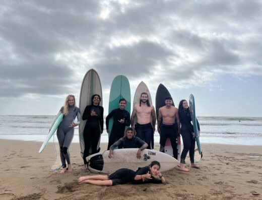 A diverse group of surfers with surfboards on the beach, smiling and posing after a surfing session at Waverii, with a cloudy sky and ocean in the background.