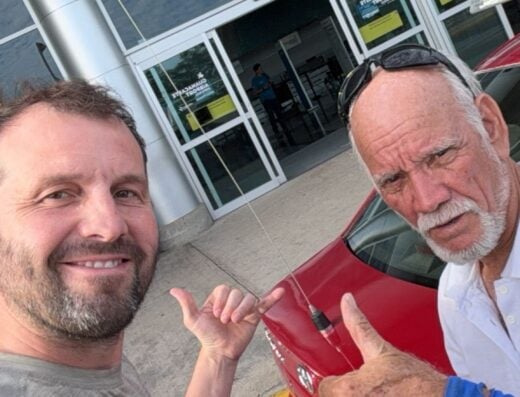 Two men taking a selfie outside an airport terminal, with a red car in the background, representing travel and adventure experiences.