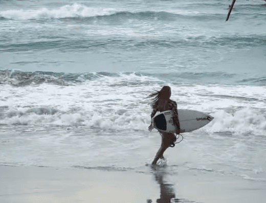 Young woman carrying surfboard walking into ocean waves at Fideluna Beach Bungalow, perfect for surfing and beach relaxation.